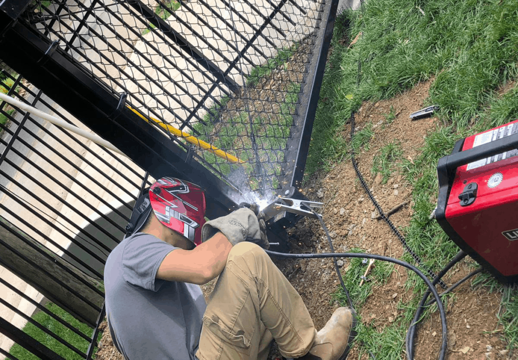 Worker welding a metal gate hinge outdoors.