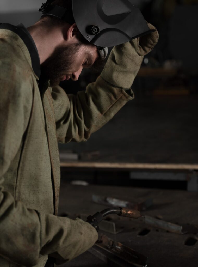 Welder in protective jacket lifting helmet while holding a welding torch over a metal workbench.